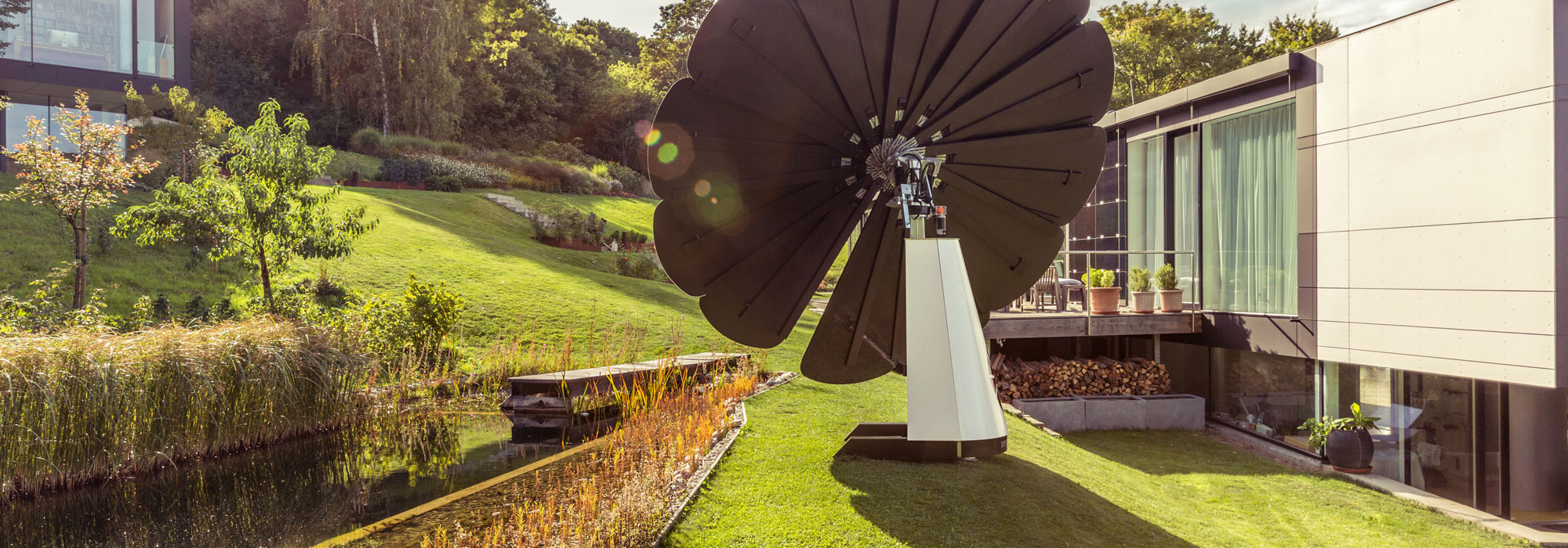 A SmartFlower Solar Panel Sits in the Yard Beside a Small Pond Between Two Houses