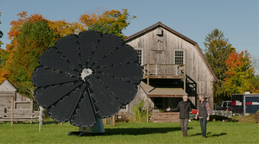 Two hosts discuss sustainable energy outside historic barn with large SmartFlower solar array