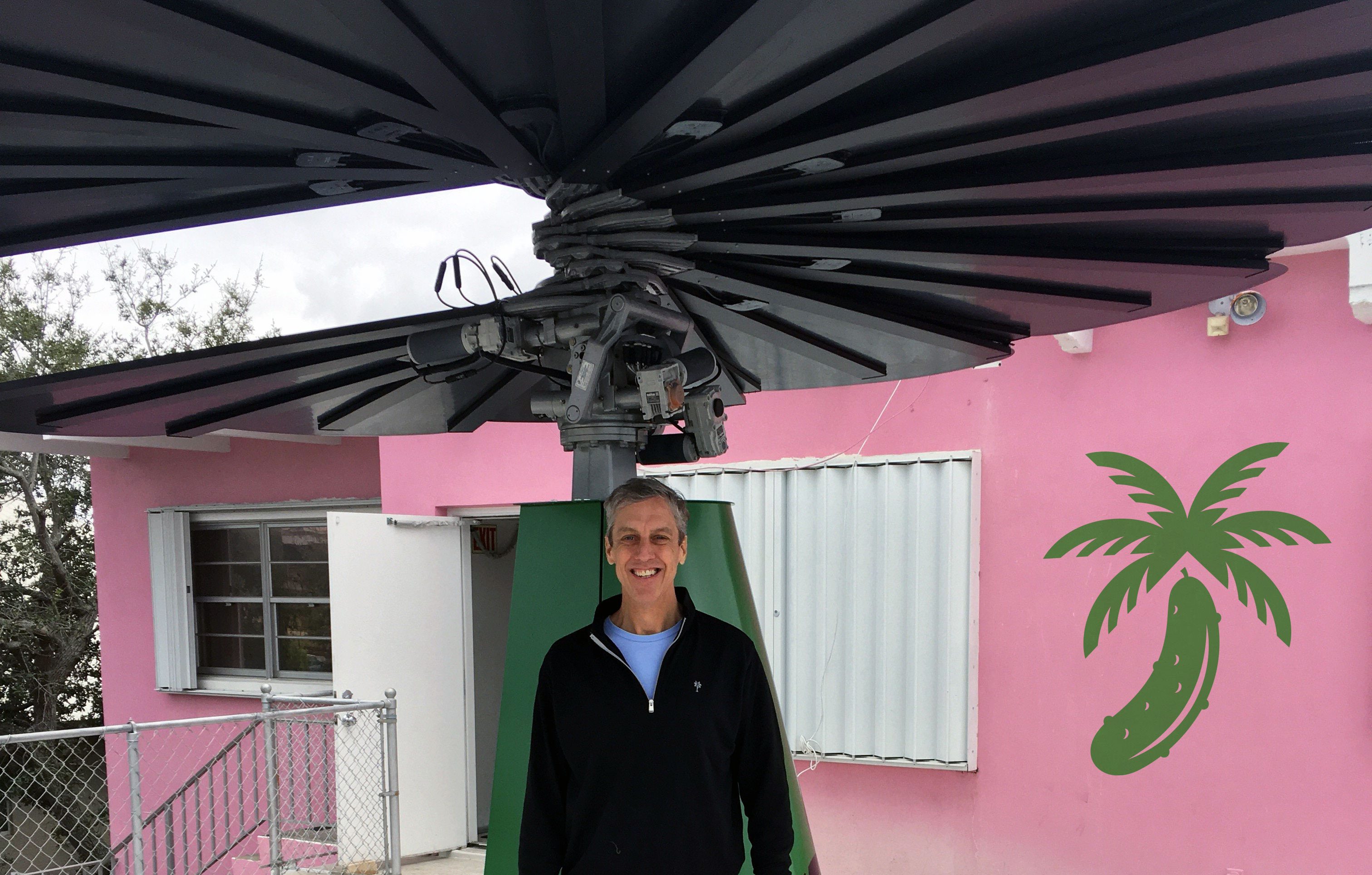 Man standing beneath Smartflower solar panel with green base in front of pink tropical building