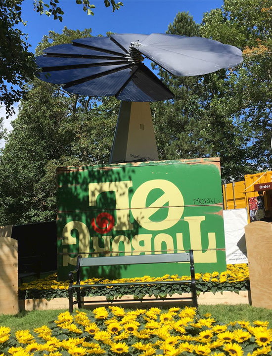 Renewable energy SmartFlower featured in garden scene with flowers, bench, and sustainability message board