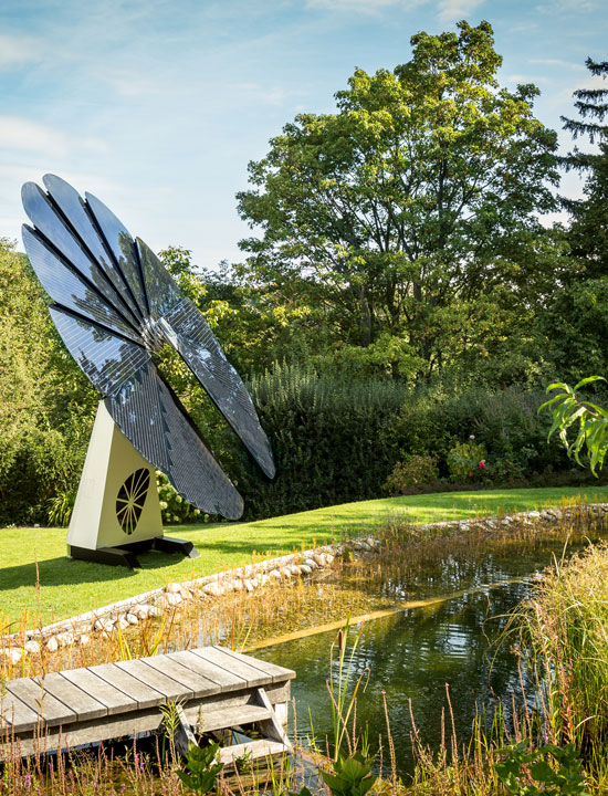 Energy-generating solar flower beside backyard pond with landscaped lawn and dense shrubbery backdrop