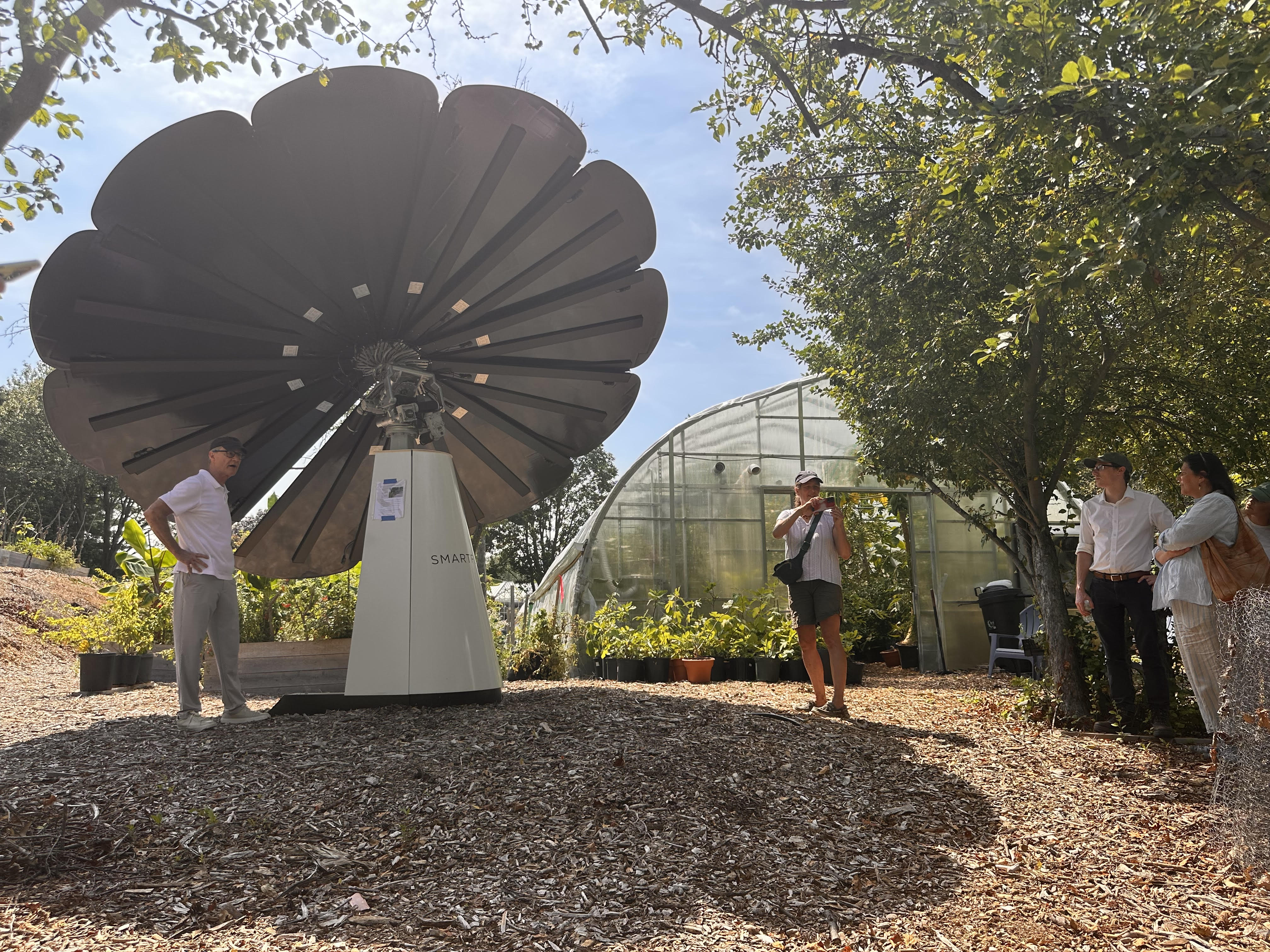 Group of people observing a large Smartflower solar panel outside a greenhouse on sunny day